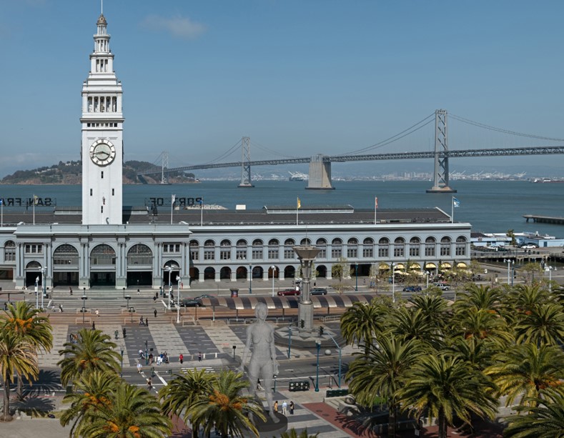 San Francisco Ferry Building with its clock tower in the foreground and the Bay Bridge stretching across the water behind it on a clear day.