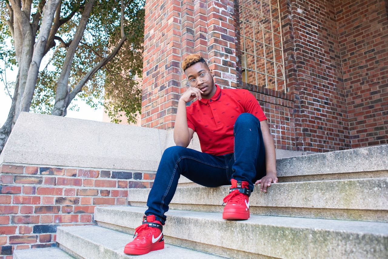 An African American trans man wearing a red polo and jeans posed in a seated position on a stone stairway