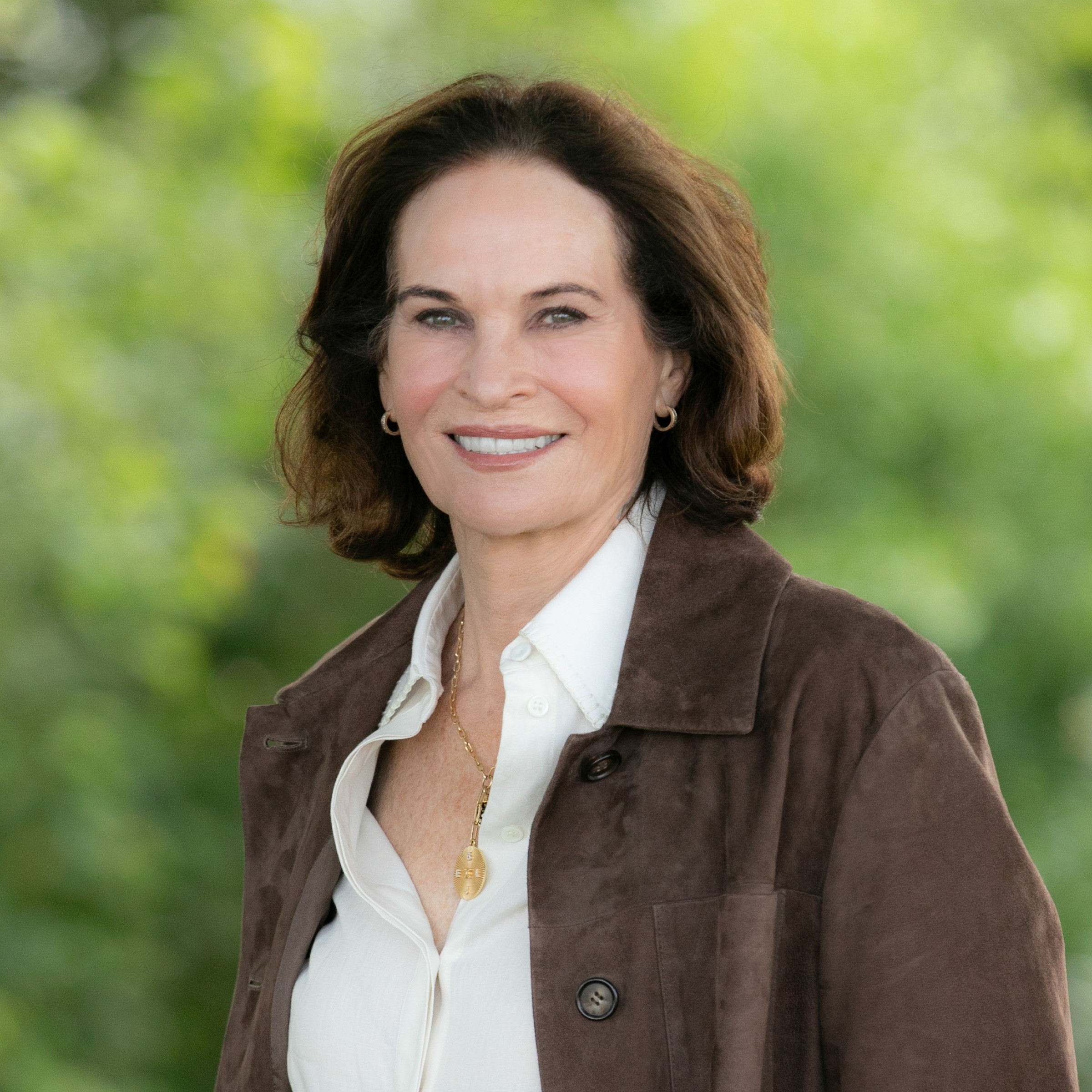 Woman with medium length brown hair wearing a white shirt and brown jacket standing in front of a green backdrop.
