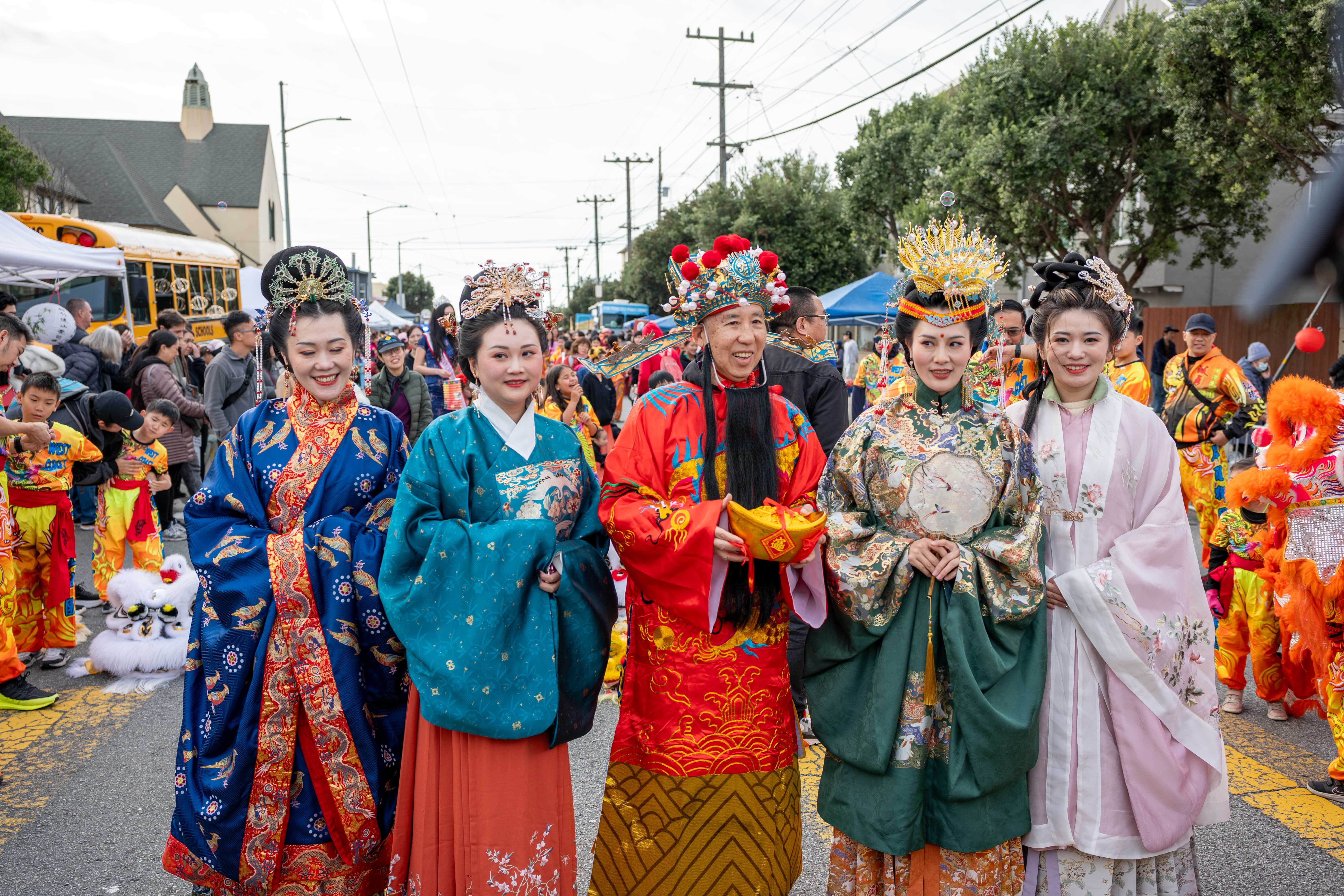 A group of five people dressed in traditional, colorful Lunar New Year costumes at a lively outdoor festival. The atmosphere is vibrant and festive.
