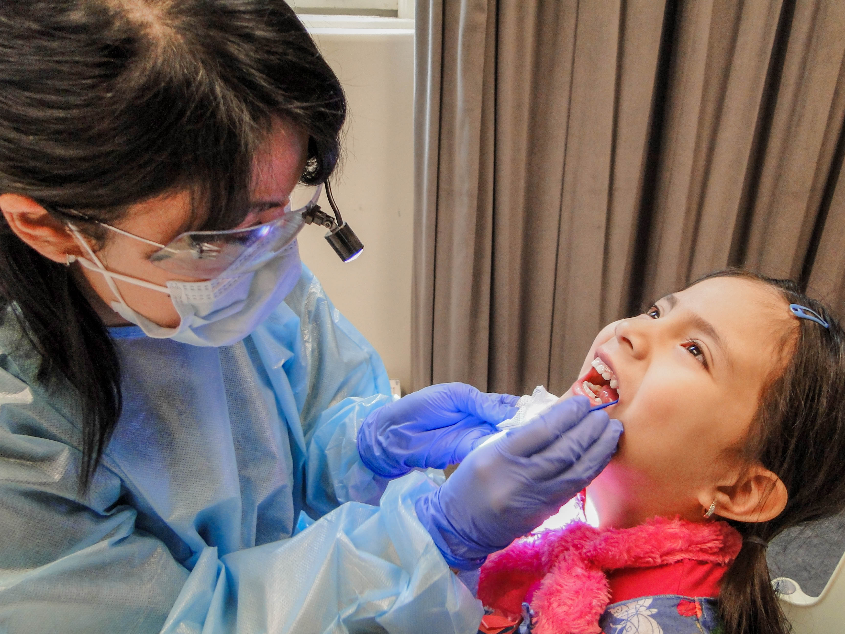 Dentist in blue protective gown and a head lamp, examines the teeth of a kindergarten age child.