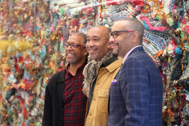 Artists Ron Moultrie Saunders, Ramekon O'Arwisters, and William Rhodes stand in front of Woven Calm, a sculptural tapestry by O'Arwisters. Photo courtesy of Nicky Woo