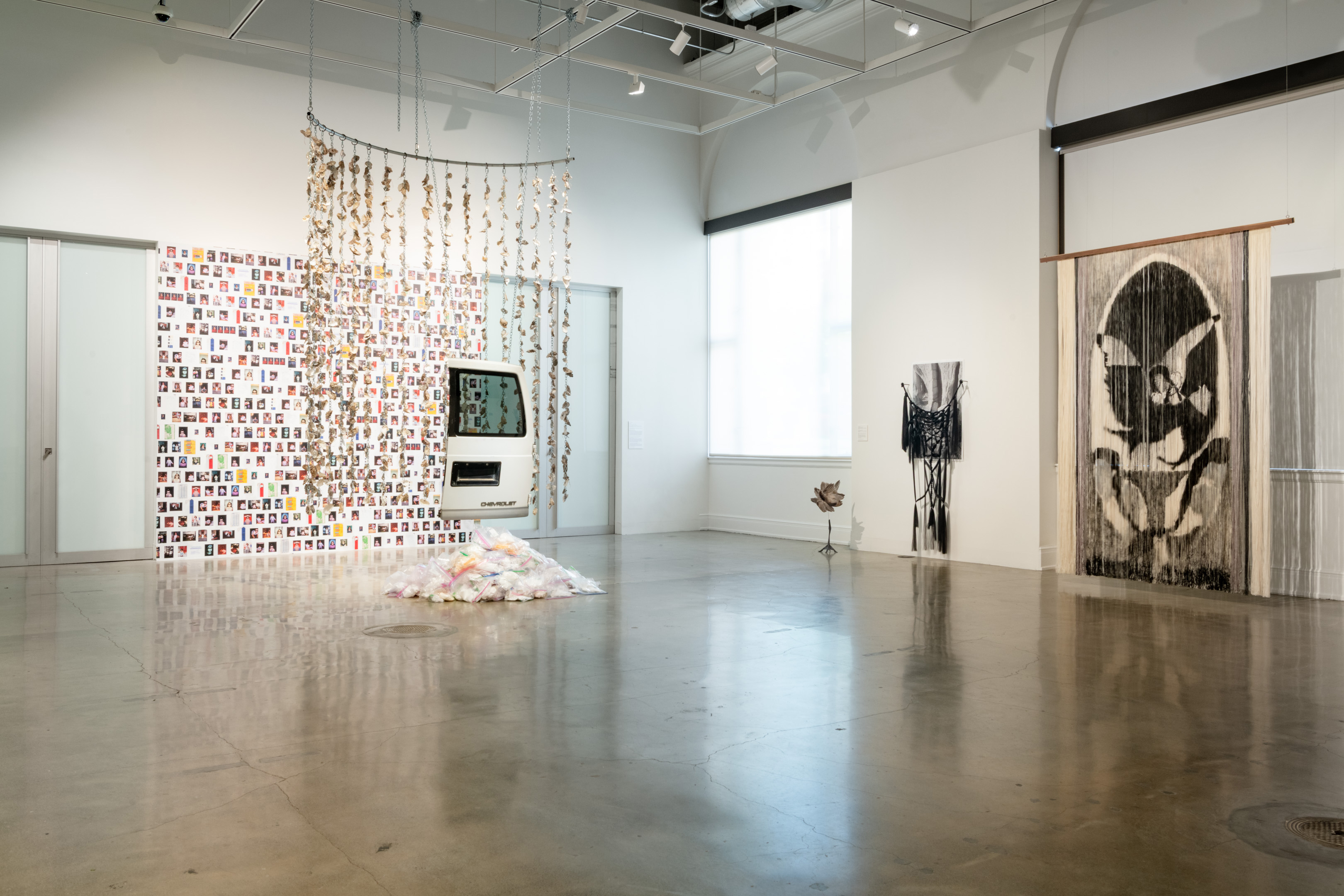 Una exposición de arte contemporáneo con una gran instalación escultórica suspendida a la izquierda y una pieza de hilo impresa y suspendida a la derecha.