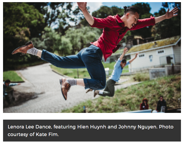 A man in a red shirt leaps midair outdoors, arms spread wide, while others jump behind him near a path and buildings.