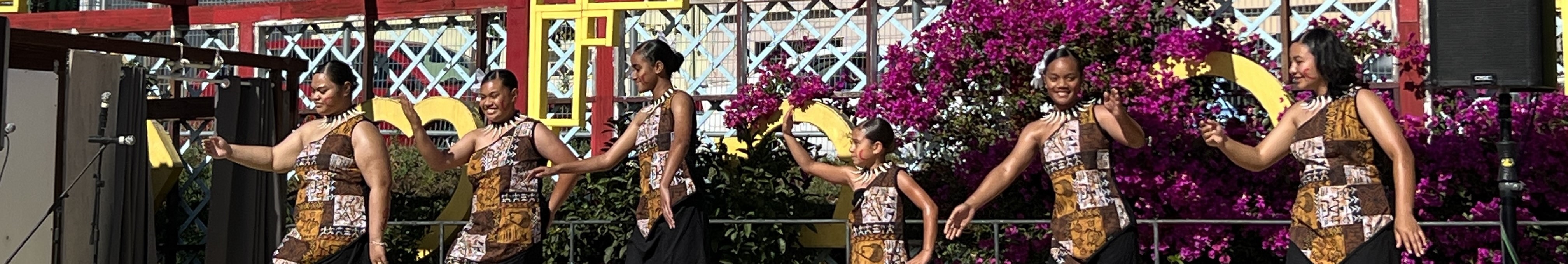 Polynesian dancers in patterned dresses perform outdoors in front of pink bougainvillea and colorful structures.