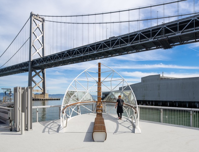 View beneath the Bay Bridge with a geometric waterfront structure and a person walking along the pier.