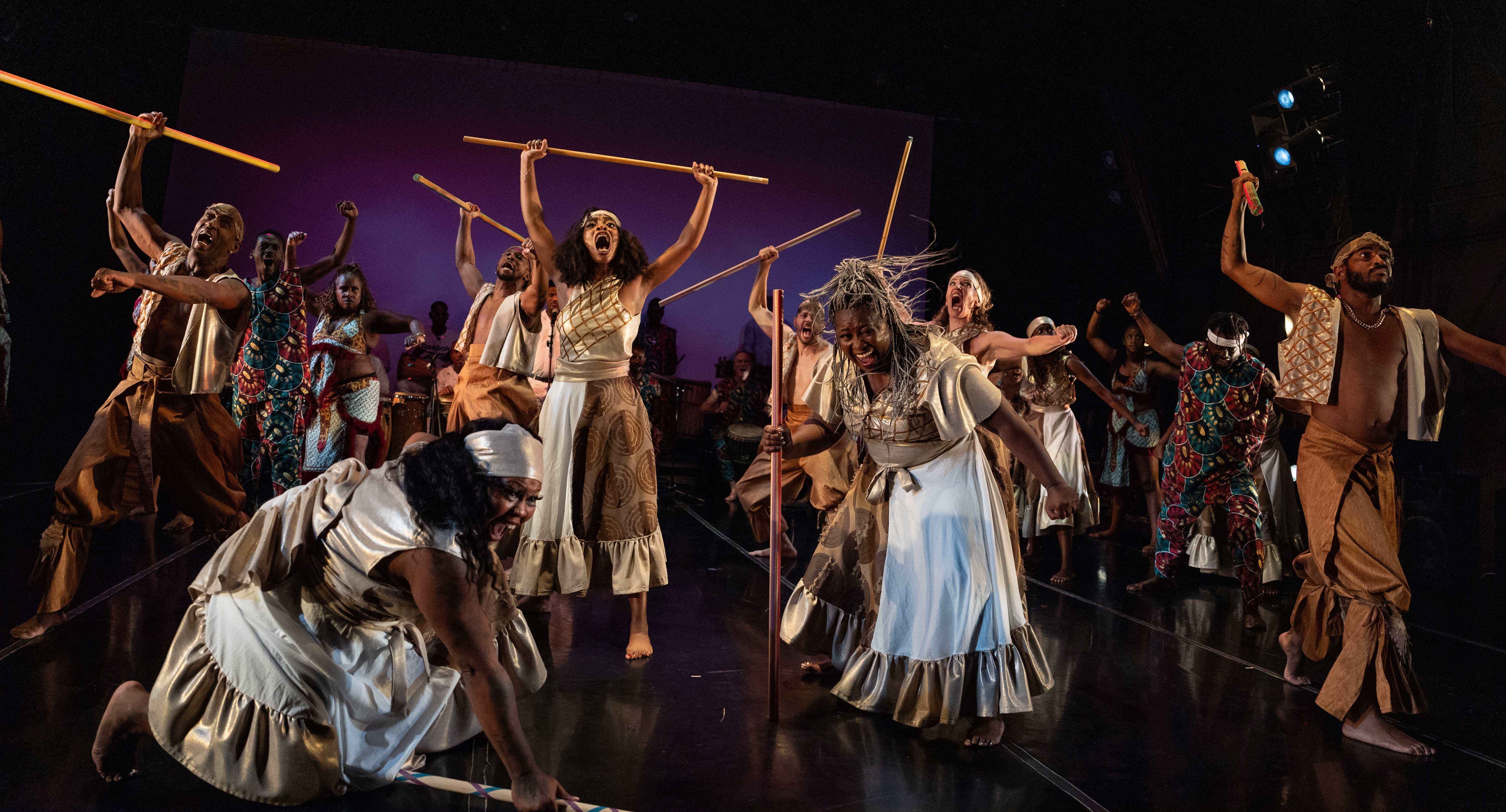 A large ensemble of dancers performs an intense, dramatic movement onstage, many holding wooden sticks raised high. The dancers wear flowing gold, white, and earth-toned costumes, with expressions of power and determination. Some crouch low while others stand tall, creating dynamic energy across the stage. Additional performers and drummers are visible in the background under warm theatrical lighting