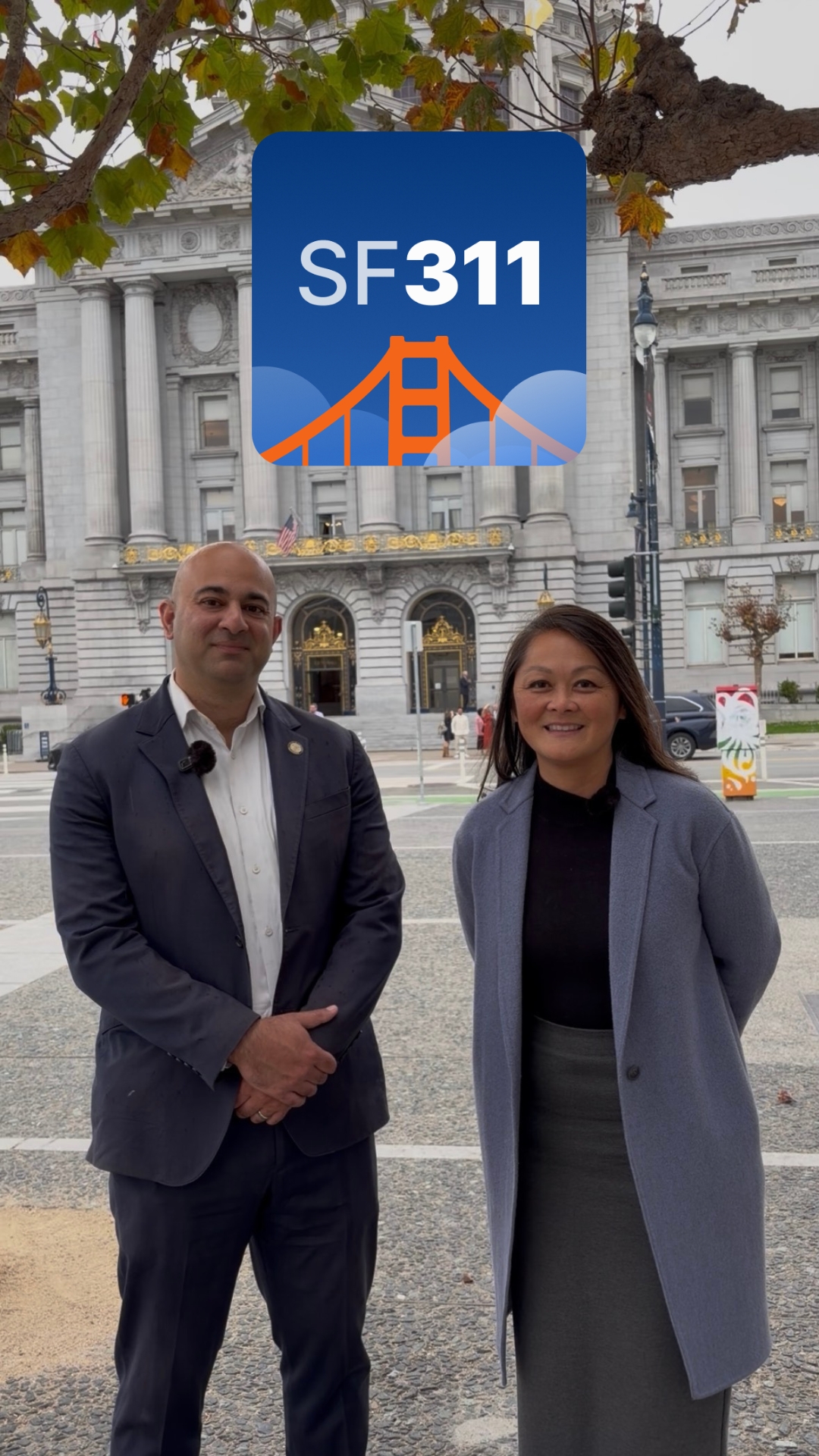 A man in a suit and a woman in a coat stand in front of City Hall with the logo for 311 above them.