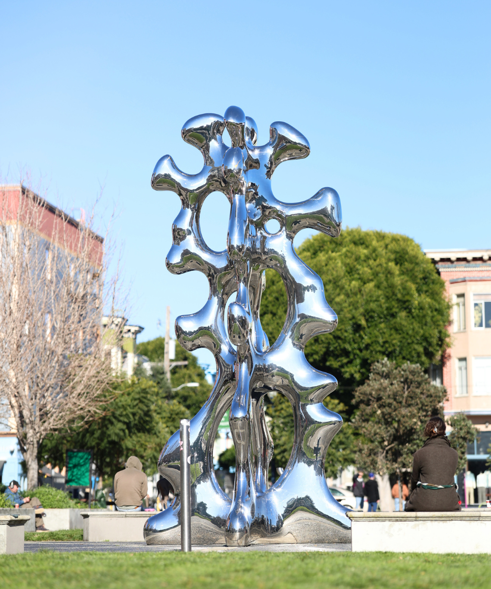Reflective abstract metal sculpture with organic shapes standing in a park, with people seated nearby and buildings in the background.