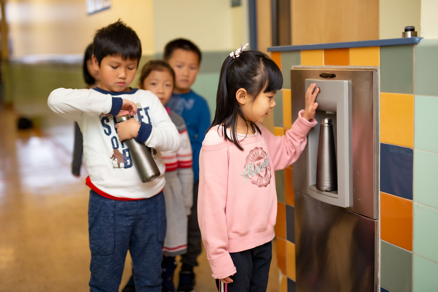A child fills a reusable water bottle at a school water station. A line of children wait behind her to fill their bottles.