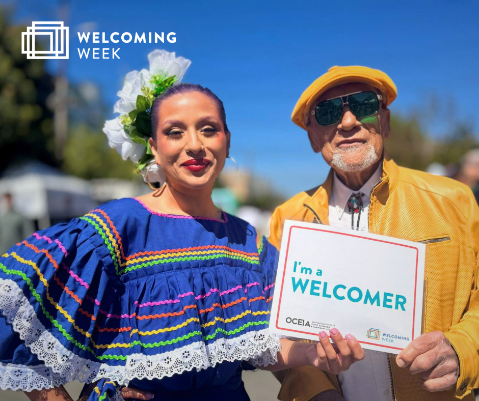 Una mujer vestida con atuendo tradicional centroamericano sonríe junto a un hombre con sombrero y chaqueta amarillos, que lleva un cartel que dice "Soy un anfitrión".