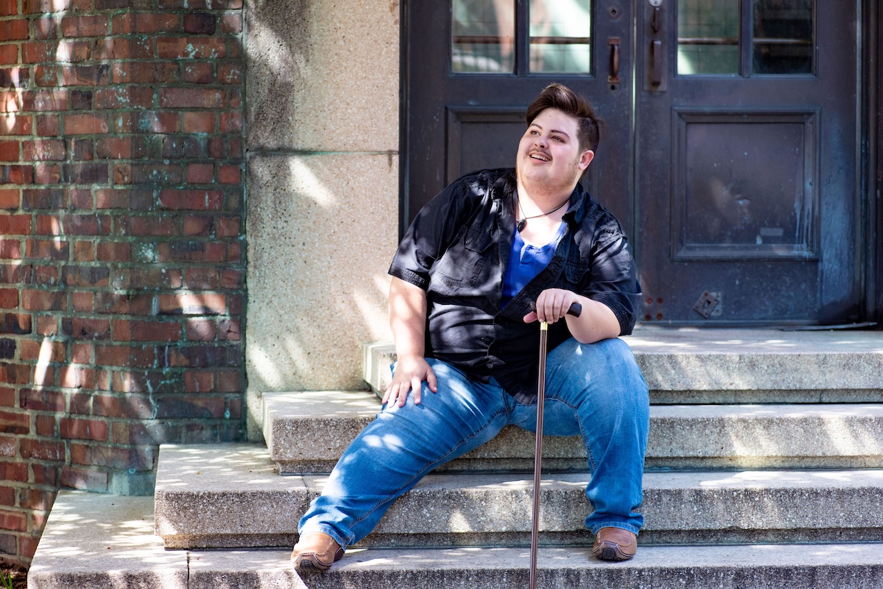 A Caucasian trans man wearing a navy button up shirt and jeans, holding a wooden cane, posed in a seated position on a stone stairway