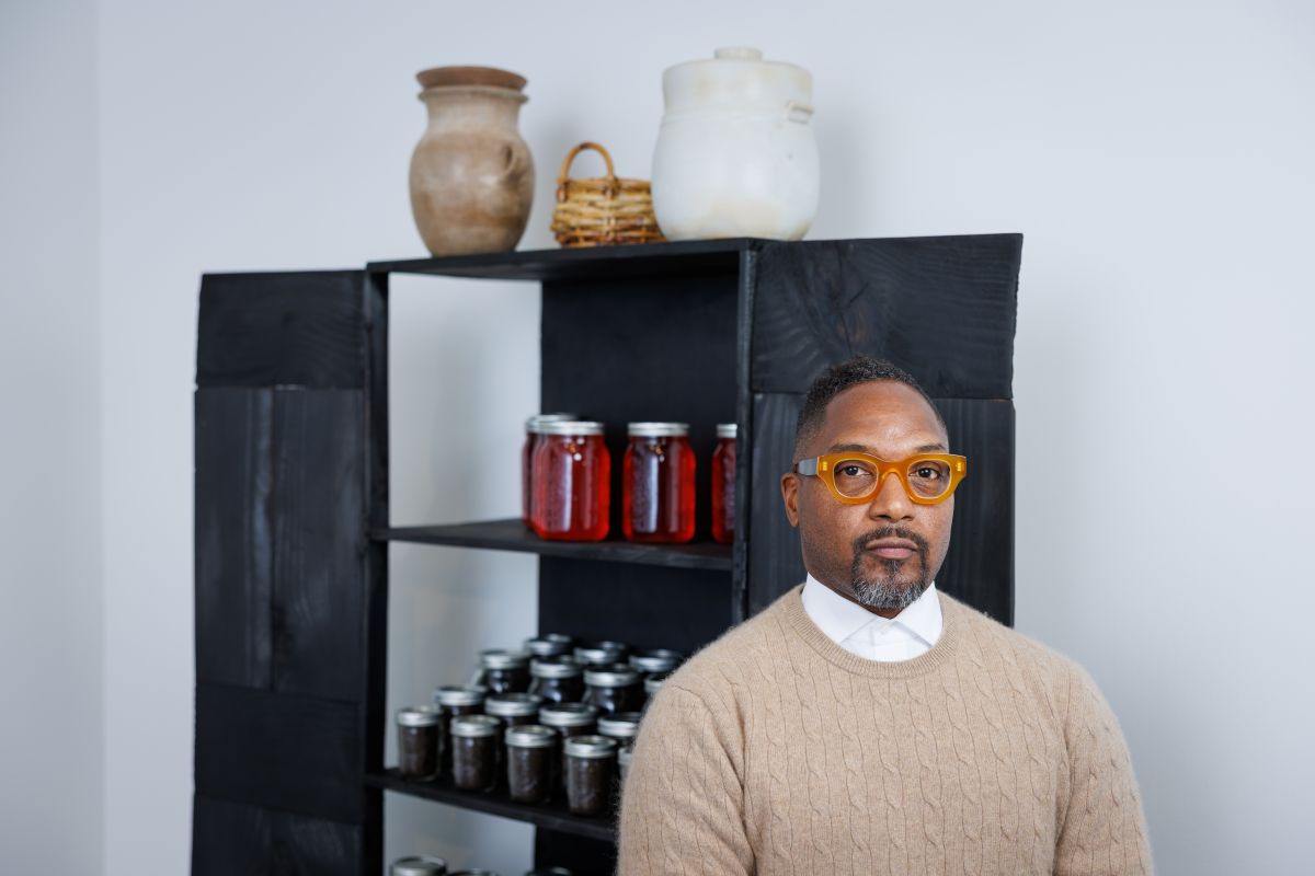 A man wearing orange colored glasses looks towards the camera. Behind him is a black cabinet filled with jars.