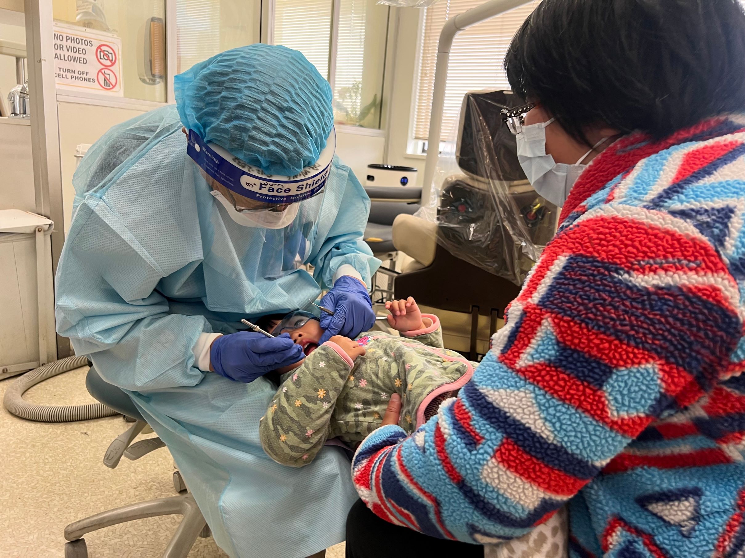 A dentist in a blue face shield examining a toddler's teeth, while the child is sitting in the parent's lap.