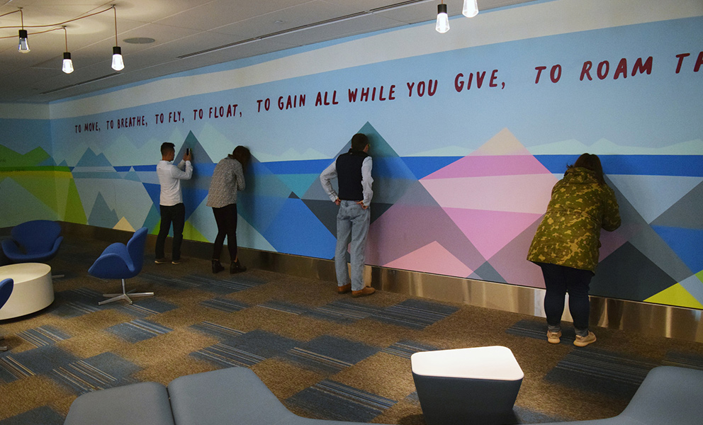 People interact with a colorful geometric wall mural featuring mountains and a quote, inside a modern indoor space.