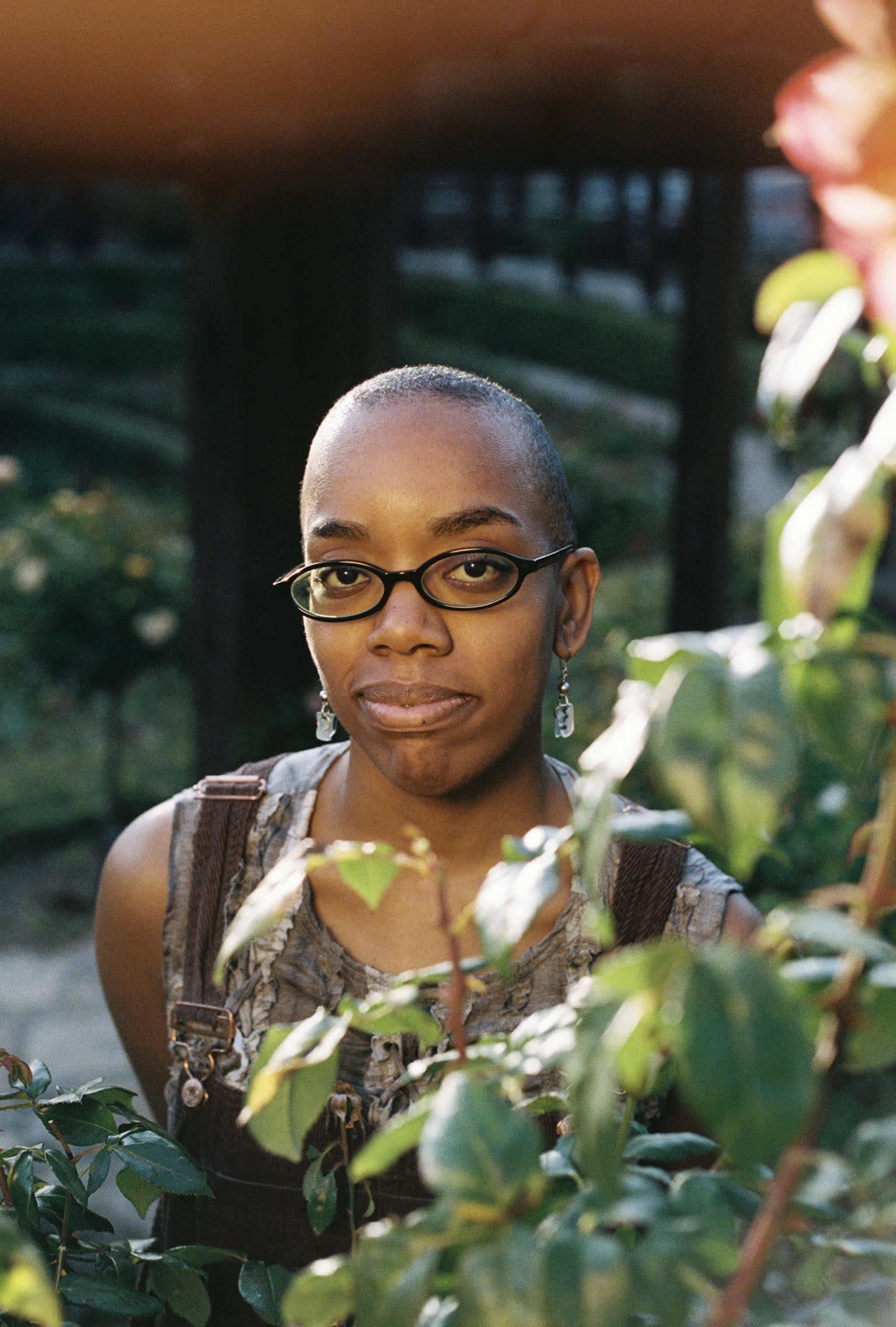 A young African-American person with a shaved head and oval glasses smirks while looking into the camera with a rose bush in the foreground.