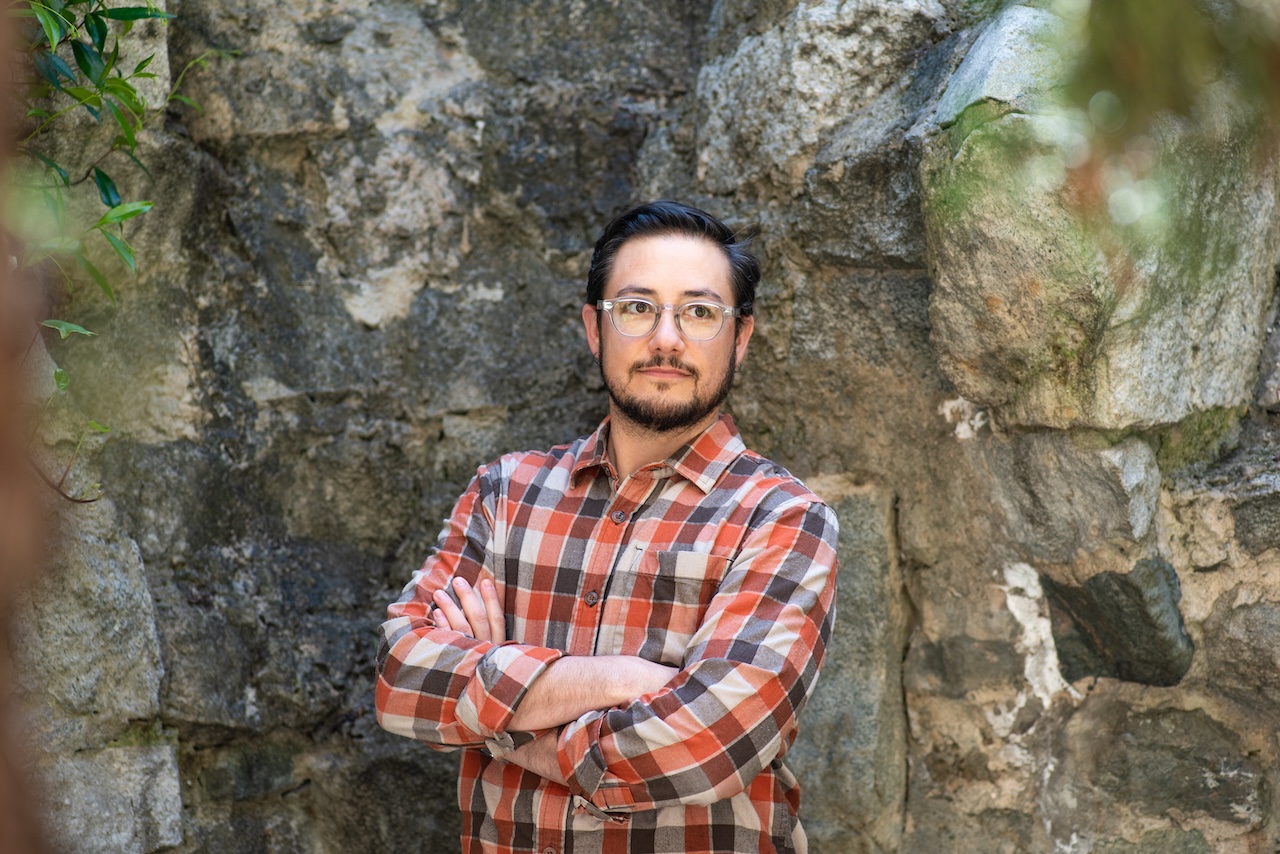 A Caucasian trans man wearing a plaid flannel posed with his arms crossed in front of a rock wall