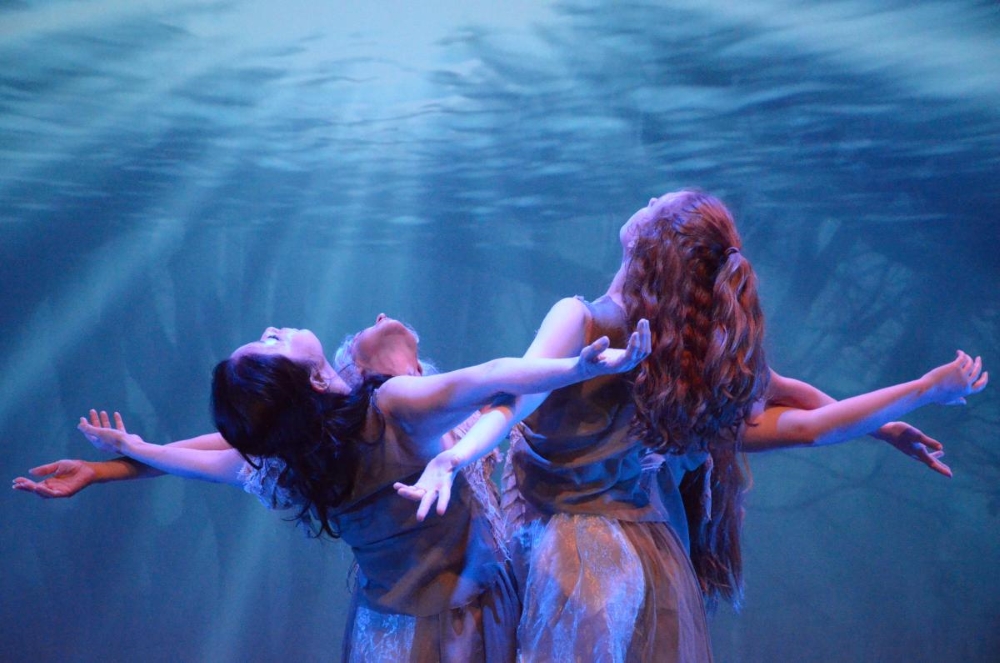Four dancers facing each other in a circle with their arms out to their sides and heads tilted back, blue light