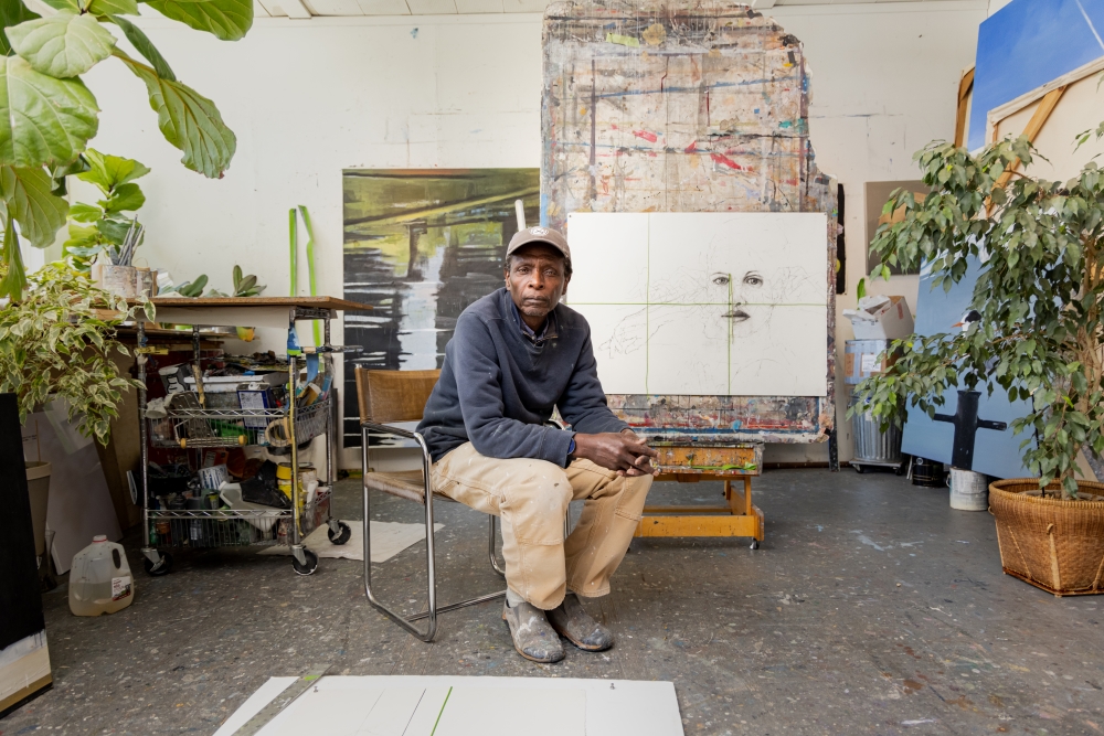 A man sits in the middle of a room in front of an easel with his painting in progress. The canvas shows the outline of a woman's face. There are art supplies and plants in the room.