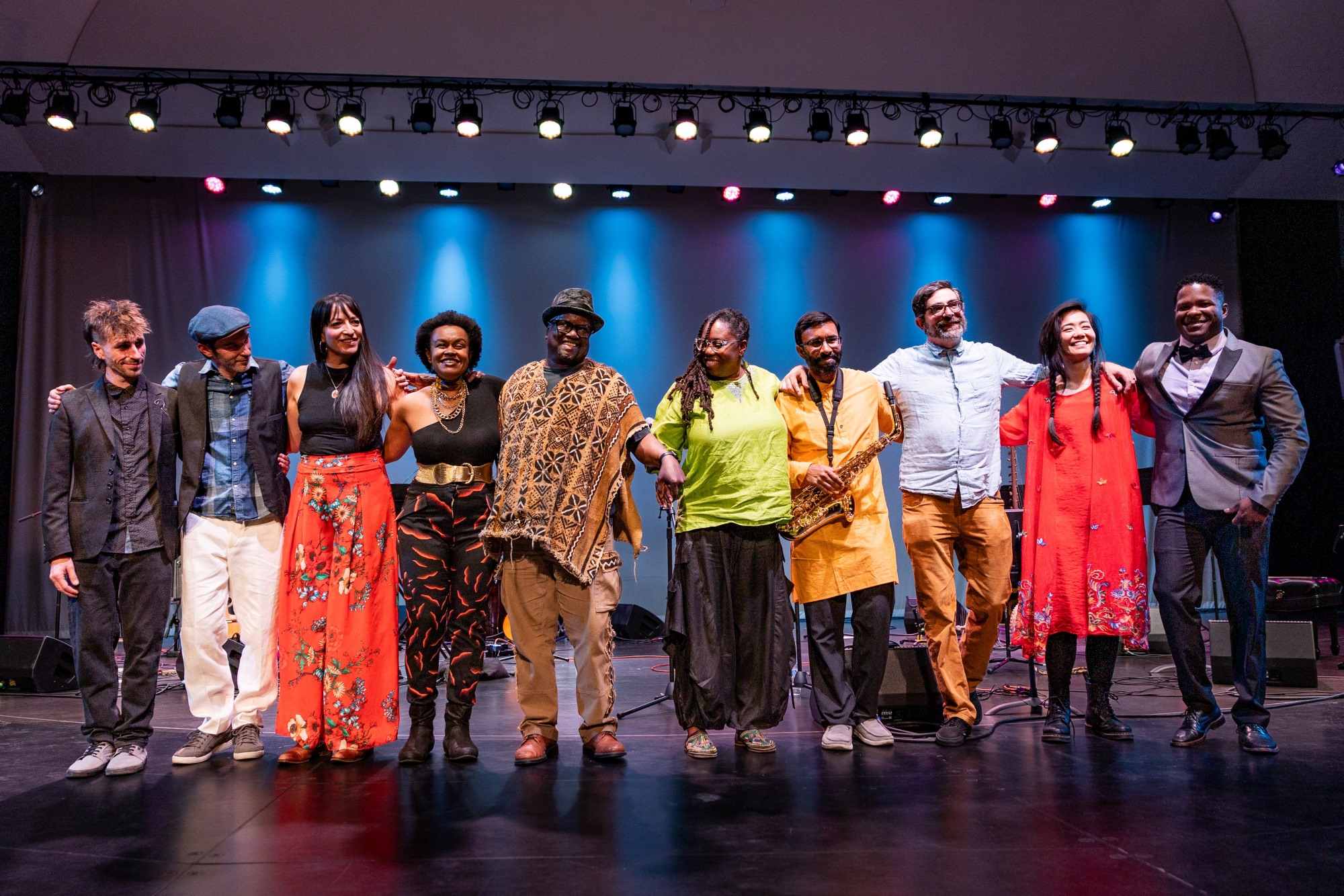Ten dancers stand in a row on a stage with white lights shining above them. They are all embracing the people next to them and are wearing colorful clothing. They are all smiling and laughing.
