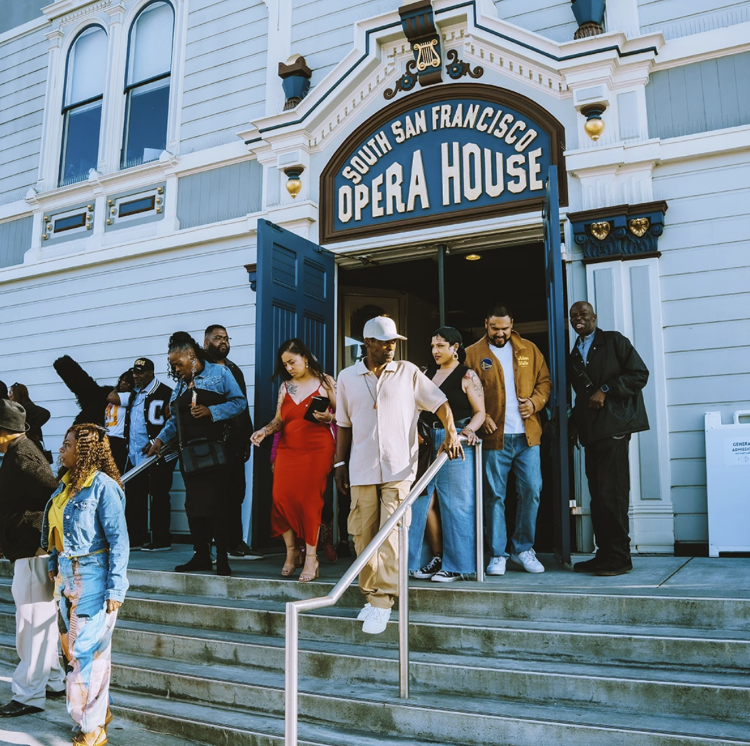 a group of people exiting a theatre door and walking down stairs, the words "South San Francisco Opera House" are on the sign above the door