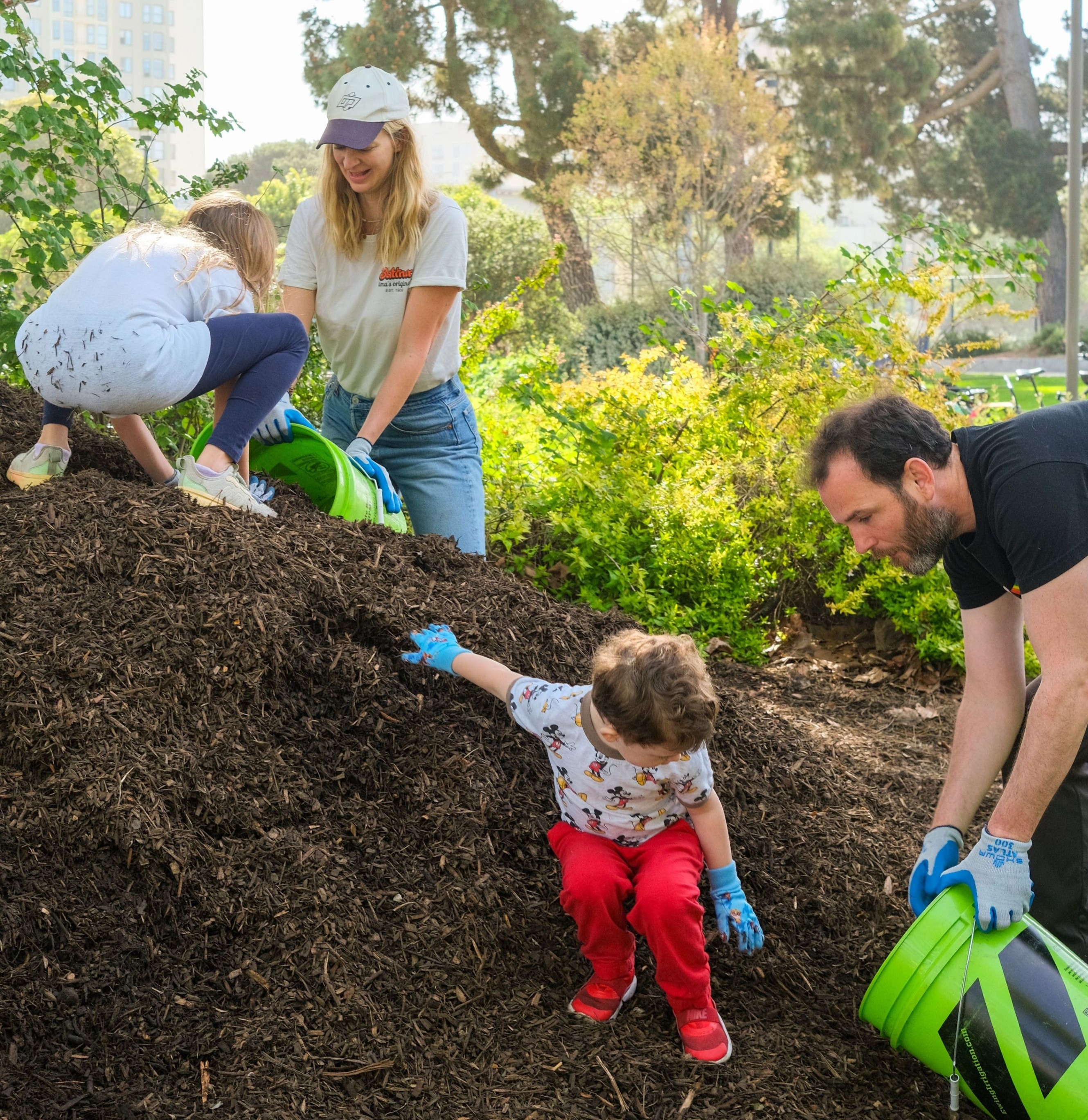 A family works together in a garden, spreading mulch. Adults and children use green buckets, enjoying sunny weather and lush greenery around them.