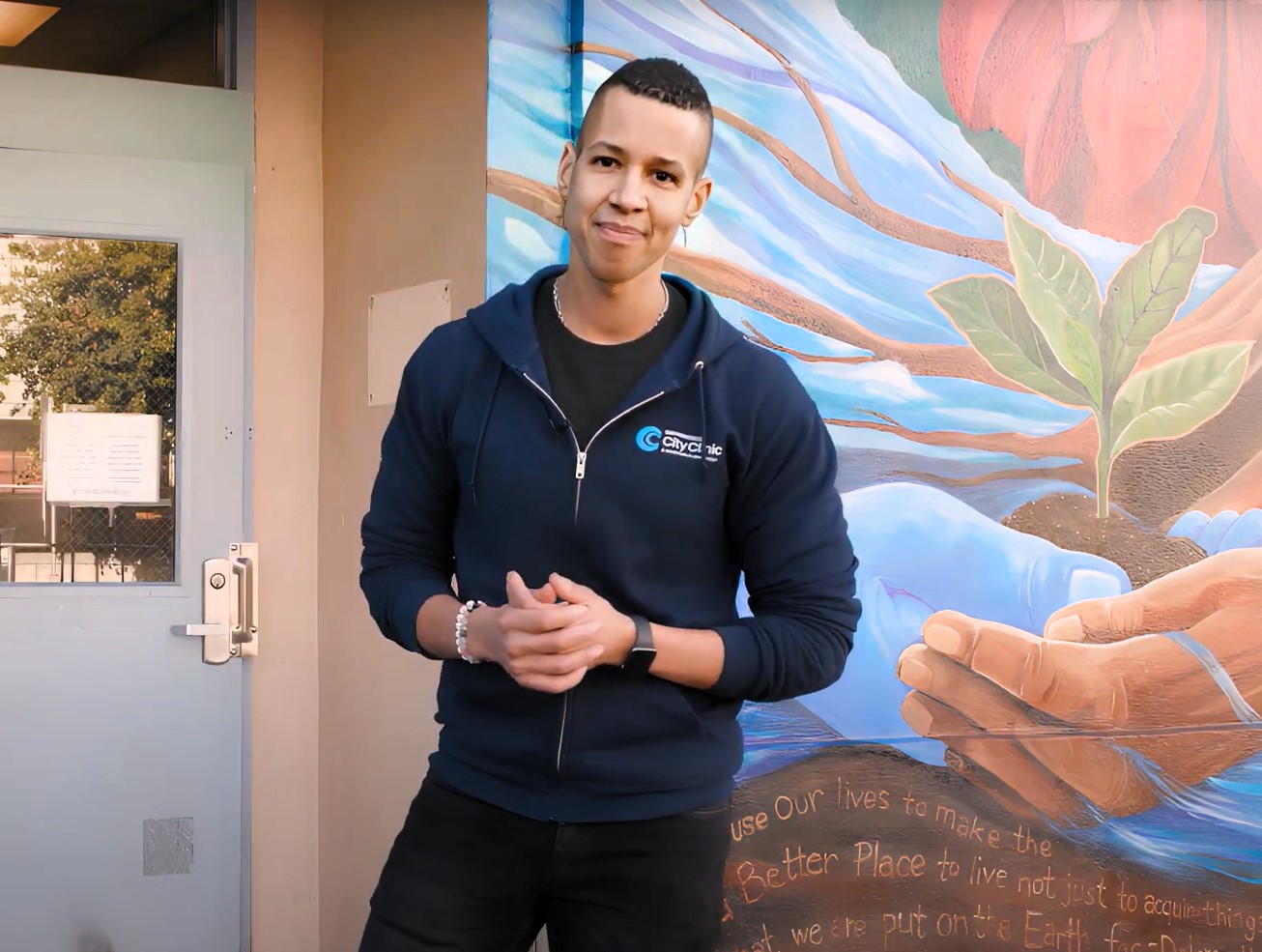 a person wearing a dark blue sweatshirt stands outside the front door of a clinic smiling